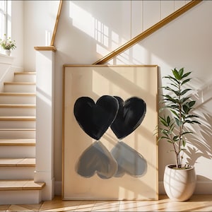 Pode incluir: Obra de arte emoldurada grande com duas formas de coração preto com seus reflexos. A obra de arte é exibida em uma entrada bem iluminada com uma escada, uma planta em vaso e uma paleta de cores neutras.