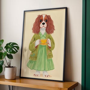 以下が含まれることがあります： 緑色のドレスとジャケットを着て、黄色のティーカップを持っているキャバリア・キング・チャールズ・スパニエルの犬が描かれた額入りのアートプリント。犬は茶色と白の毛並みで、クリーム色の背景に黒いフレームで描かれています。