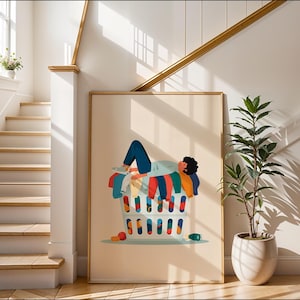 May include: A framed art print shows a person relaxing in a laundry basket filled with colourful clothes. The illustration is set against a neutral background, with a staircase and a potted plant in the scene.