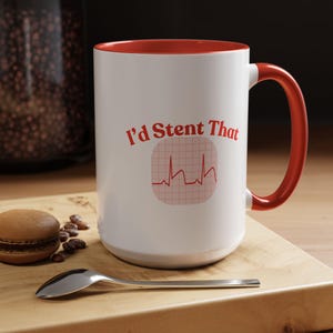 Può includere: Tazza in ceramica bianca con interno e manico rossi. La tazza presenta il testo "I'd Stent That" in rosso, insieme a un grafico a forma di battito cardiaco ECG rosso. La tazza è su una superficie di legno con un cucchiaio e un macaron.