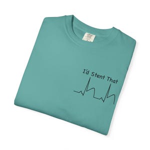 Op de afbeelding: Een blauwgroen T-shirt met ronde hals en een zwarte opdruk aan de rechtervoorzijde. De afbeelding toont een hartslaglijn en de tekst "I'd Stent That" in een eenvoudig zwart lettertype. Het label aan de binnenzijde van de hals is zichtbaar.