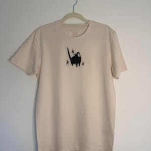 May include: A cream-coloured short-sleeve t-shirt featuring a centred black ink-style illustration of a cat with its mouth open, surrounded by three small four-pointed star shapes. The shirt has a crew neckline and is displayed on a hanger.