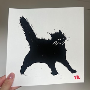 May include: A monochrome print featuring a black cat with an arched back and a raised tail. The cat is depicted mid-stride, with its mouth open and whiskers showing. The artist's initials, "rk", are in red in the bottom right corner.