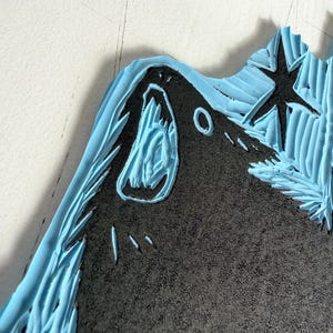 May include: Close-up of a carved linoleum block with a black design and light blue border. The design includes a star, a mountain, and a stylized animal head. The linoleum has a textured surface.