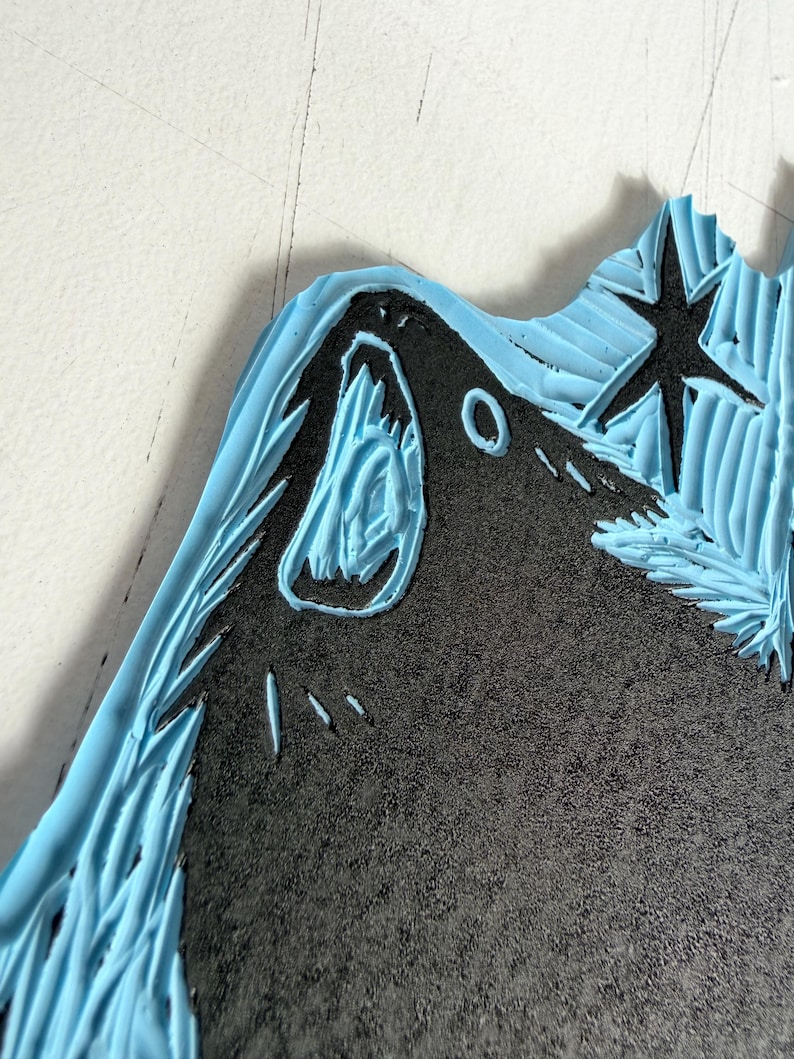 May include: Close-up of a carved linoleum block with a black design and light blue border. The design includes a star, a mountain, and a stylized animal head. The linoleum has a textured surface.