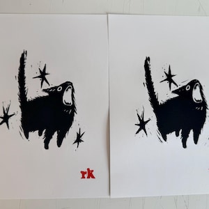 May include: Two identical black and white art prints. Each print features a stylized black cat with an open mouth, surrounded by star shapes. The artist's initials, "rk", are in red at the bottom.