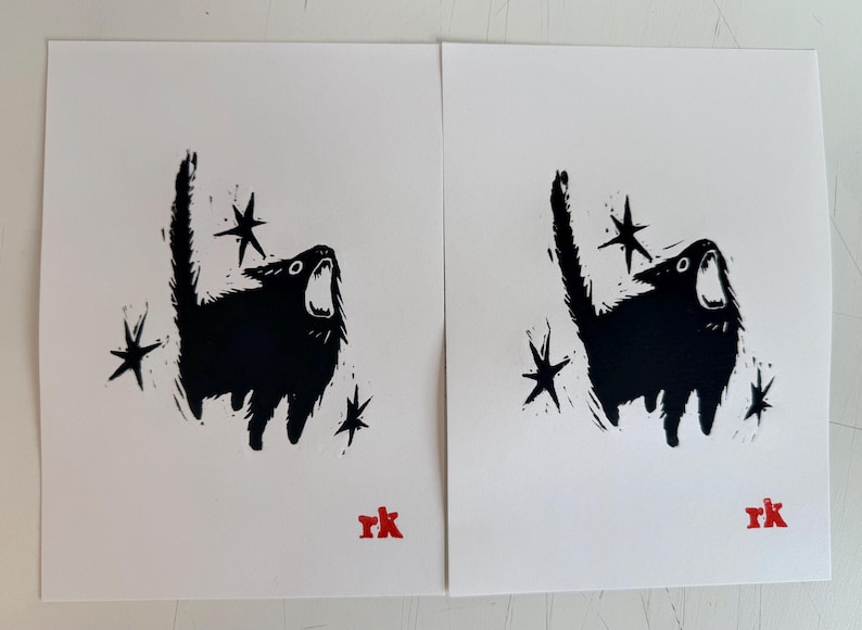 May include: Two identical black and white art prints. Each print features a stylized black cat with an open mouth, surrounded by star shapes. The artist's initials, "rk", are in red at the bottom.