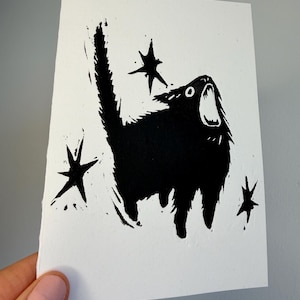 Peut inclure: Estampe en linogravure noir et blanc d'un chat avec la bouche ouverte, hurlant ou miaulant. Le chat est entouré de formes d'étoiles. L'estampe est sur une carte rectangulaire blanche.