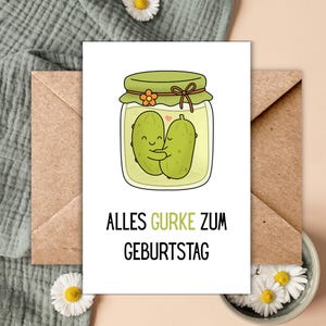 Può includere: Biglietto di auguri con un'illustrazione a cartoni animati di due cetrioli sottaceto che si abbracciano in un barattolo, con il testo tedesco "Alles Gurke Zum Geburtstag". Il biglietto è su sfondo bianco, con una busta marrone e margherite.