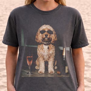 May include: A charcoal gray crew neck t-shirt featuring a graphic print of a light brown curly-haired dog wearing black sunglasses. The dog is positioned between a tall glass of pink liquid and a dark bottle on a flat surface.
