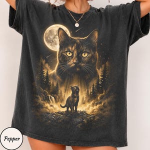 May include: A charcoal gray short-sleeve t-shirt featuring a graphic print of a large cat face above a smaller cat standing on a rocky landscape with a forest, a waterfall, and a full moon in the background. The word Pepper is printed in a circle.