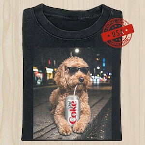 May include: A black crew neck t-shirt featuring a graphic of a golden-colored dog wearing black sunglasses and holding a silver Diet Coke can in its mouth. The background shows a blurred city street at night with bokeh lights and a red USA stamp.