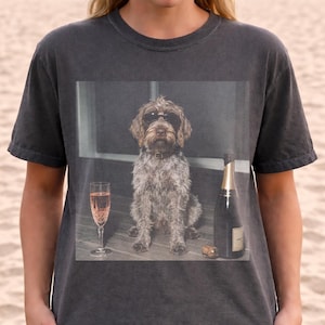 May include: A charcoal gray t-shirt featuring a graphic print of a wire-haired dog wearing dark sunglasses, sitting between a tall glass of pink sparkling wine and a dark glass bottle of champagne with a cork resting on the surface.