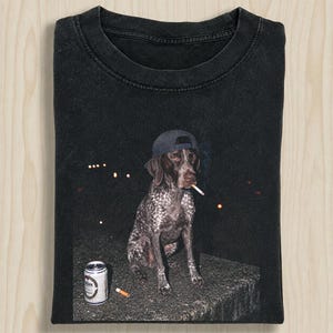 May include: A black crew neck t-shirt featuring a graphic print of a spotted dog wearing a baseball cap, holding a cigarette in its mouth, and sitting next to a silver beverage can and a discarded cigarette butt on a dark surface.