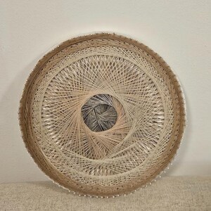 May include: A circular wall hanging made of string, with a brown and beige color scheme. The string is woven in a circular pattern, creating a spiral effect. The center of the hanging is a smaller circle of string, with a darker brown color.