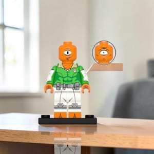 Puede incluir: Una minifigura de plástico con cabeza naranja de cíclope, camisa verde con detalles blancos y pantalones blancos. La figura se sostiene sobre una base negra, con una cabeza de cíclope naranja adicional en un pequeño estante lateral.