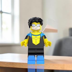 Puede incluir: Una minifigura de plástico con cabello negro, cara amarilla con gafas blancas y un traje azul y negro con un emblema amarillo en el pecho. La figura tiene brazos amarillos, manos negras, caderas negras y piernas azules en postura neutral.