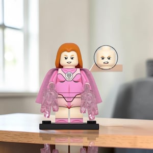 Könnte beinhalten: Eine Kunststoff-Minifigur mit orangefarbenem Haar, die einen rosa Body mit einem weißen kreisförmigen Emblem auf der Brust und einen passenden rosa Umhang trägt. Die Figur hat durchscheinende rosa Hand-Accessoires und einen Ersatzkopf mit wütendem Gesichtsausdruck.
