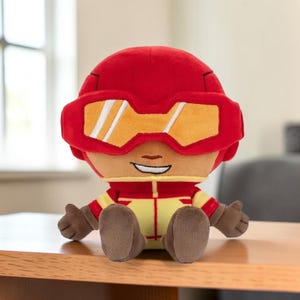May include: A plush toy character wearing a red helmet with large yellow goggles, a red and yellow suit, and brown gloves and boots. The character has a smiling expression with visible teeth and is depicted in a seated position with arms extended.