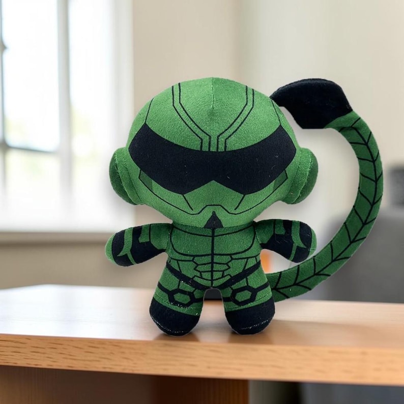 May include: A green and black plush toy featuring a character with a rounded head, a visor-like face mask, and a long, segmented tail that curves upwards. The body is decorated with dark lines resembling armour plating on the chest and limbs.