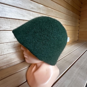 Może przedstawiać: Ciemnozielony, puszysty kapelusz w stylu cloche. Kapelusz wykonany jest z miękkiego, fakturowanego materiału.