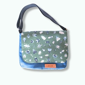 May include: A blue and green messenger bag with a flap featuring a pattern of various insects in white and blue. The bag has a blue denim-like base and a dark gray adjustable strap. A small brown tag reads "JACOB".