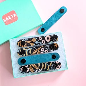 May include: A set of four button straps, two with a floral pattern and two solid teal, displayed in a teal box. The box has a label that reads "LAETA HANDMADE GOODS". The straps are made of leather and have black button closures.