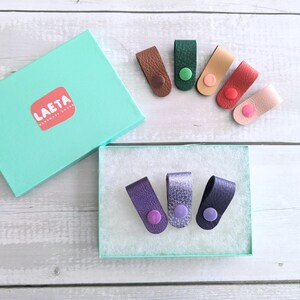May include: A selection of leather cord tidies in shades of brown, green, beige, red, and pink, each with a matching press stud. The organizers are presented in a mint green box and a clear box with white filling. The boxes have the text "LAETA HANDMADE GOODS".