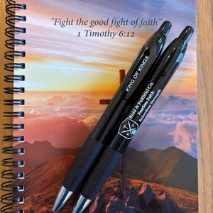 May include: A black spiral bound notebook with a sunset and mountain scene on the cover. The notebook has the text "Fight the good fight of faith" and "1 Timothy 6:12" printed on the cover. Two black pens with the words "King of Kings" and "Bold & Faithful Co." are on top of the notebook.