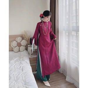 May include: A magenta Áo dài dress, a traditional Vietnamese outfit, with long sleeves and a high collar. The dress has a side slit showing green trousers. A pearl necklace and a red flower are visible. The outfit is completed with white shoes.