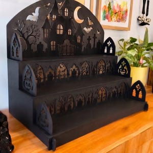 Peut inclure: Une étagère de présentation noire à trois niveaux avec des motifs découpés au laser représentant une maison hantée, un croissant de lune, des chauves-souris, des arbres et des pierres tombales. Les panneaux latéraux présentent des arches gothiques.