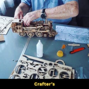May include: A wooden model train is being assembled on a table. The train features intricate details, including wheels, gears, and a cabin. A bottle of glue and other tools are nearby. The text "Crafter's" is at the bottom of the image.