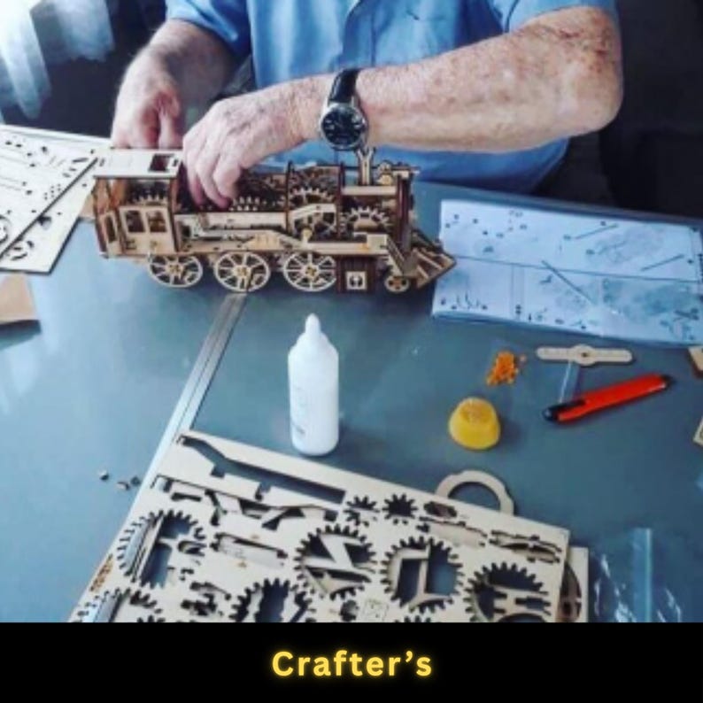 May include: A wooden model train is being assembled on a table. The train features intricate details, including wheels, gears, and a cabin. A bottle of glue and other tools are nearby. The text "Crafter's" is at the bottom of the image.