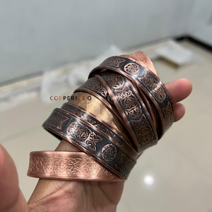 May include: A collection of copper bracelets with intricate Celtic knot designs. The bracelets have a dark, oxidized finish on the engraved areas, contrasting with the polished copper. The bracelets are stacked, showcasing the detailed patterns.