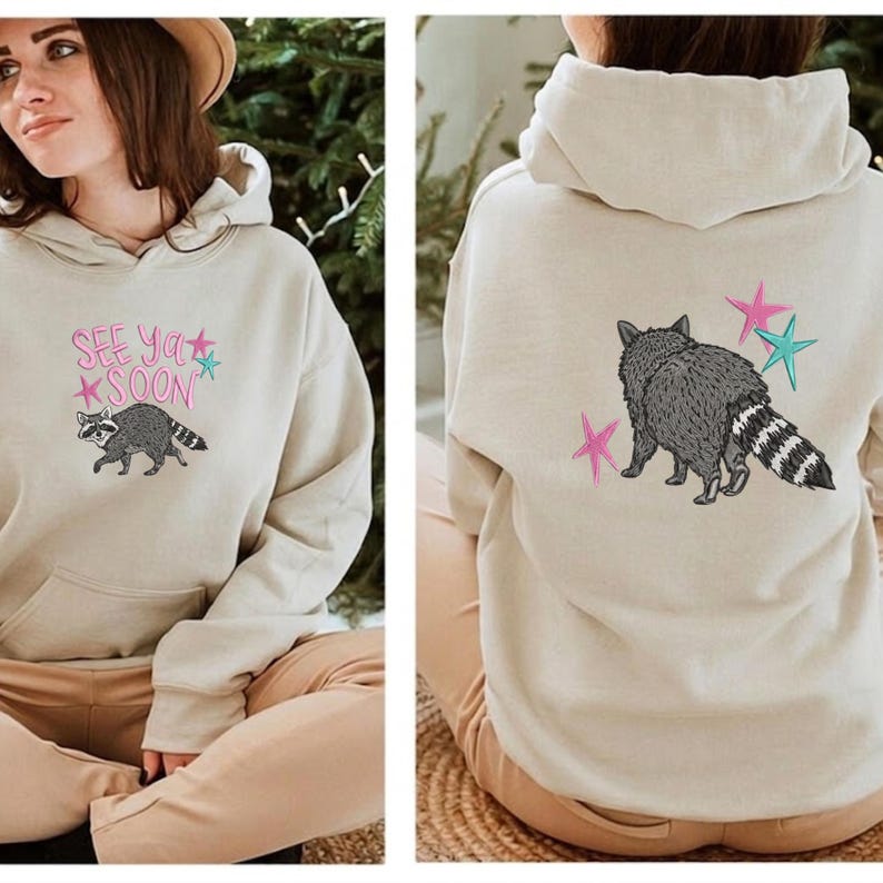 K&ouml;nnte beinhalten: Beigefarbener Kapuzenpullover mit einem Waschb&auml;r-Motiv. Auf der Vorderseite des Hoodies steht der Text "See Ya Soon" mit einer Waschb&auml;r-Illustration. Auf der R&uuml;ckseite des Hoodies ist ein Waschb&auml;r mit rosa und t&uuml;rkisfarbenen Sternen abgebildet.