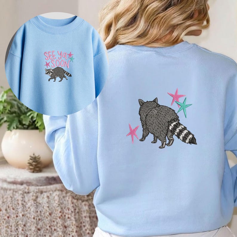 K&ouml;nnte beinhalten: Hellblaues Sweatshirt mit einem Waschb&auml;ren-Design und dem Text "See Ya Soon" und Sternen. Auf der R&uuml;ckseite des Sweatshirts ist ein Waschb&auml;r mit rosa und t&uuml;rkisfarbenen Sternen abgebildet. Der Waschb&auml;r hat einen grauen K&ouml;rper und einen gestreiften Schwanz.