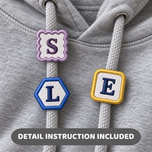 May include: A heather grey hoodie featuring two thick drawstrings. Three embroidered patches are attached to the cords: a lavender scalloped square with the letter S, a blue hexagon with the letter L, and a yellow square with the letter E.