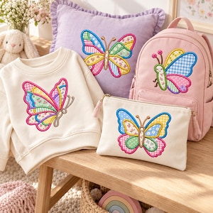 Puede incluir: Una sudadera color crema, un cojín lavanda con bordes con volantes, una mochila rosa y un estuche con cremallera color crema. Cada artículo presenta una mariposa bordada colorida con patrones estilo patchwork en tonos rosa, azul, amarillo y verde.