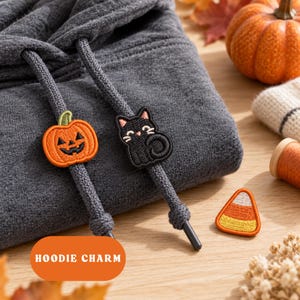 Puede incluir: Una sudadera con capucha gris oscuro que presenta dos dijes bordados en los cordones: una calabaza naranja de Halloween y un gato negro. Un parche de caramelo de maíz se encuentra cerca. El texto HOODIE CHARM aparece en un rectángulo naranja.