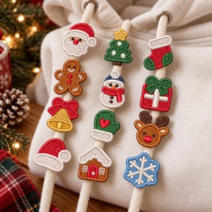 Puede incluir: Una sudadera con capucha color crema que presenta tres cordones decorados con parches bordados de temática navideña, incluyendo un Santa Claus, hombre de jengibre, campana, gorro, árbol, muñeco de nieve, manopla, casa, calcetín, regalo, reno y un copo de nieve.