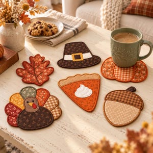 Puede incluir: Un juego de seis posavasos de tela con formas temáticas de otoño que incluyen un pavo, una calabaza, una bellota, un trozo de tarta con nata, un sombrero de peregrino y una hoja de roble, decorados con varios patrones marrones, naranjas y crema.