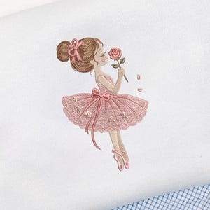 Puede incluir: Un diseño bordado sobre tela blanca que muestra una figura con cabello castaño recogido en un moño, vistiendo un tutú rosa con un lazo y detalles de encaje. La figura sostiene una rosa rosa, con dos pequeños pétalos flotantes, y lleva zapatillas de ballet rosas.
