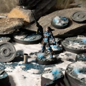 Puede incluir: Figura en miniatura de un soldado futurista con armadura azul y negra, sosteniendo un rifle. La figura se encuentra sobre una base detallada con nieve y mechones azul verdosos. Varias otras bases y rocas rodean la figura, creando una escena de diorama.