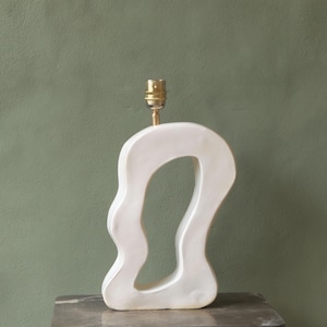 Könnte beinhalten: Eine weiße Tischlampe aus Keramik mit einer abstrakten, organischen Form. Die Lampe hat eine große, unregelmäßige Öffnung in der Mitte und eine Lampenfassung aus Messing. Sie steht auf einer dunklen Oberfläche vor einem gedämpften grünen Hintergrund.