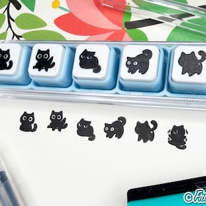 May include: A set of six square rubber stamps with light blue plastic bases, each featuring a unique black cat illustration. The stamps are housed in a clear plastic case, with corresponding black ink impressions of each cat displayed below.