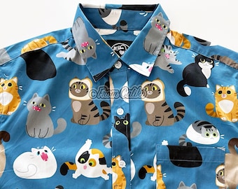 May include: A blue button-down shirt with a repeating pattern of cats in various poses and colours. The cats are wearing different expressions and some are holding objects like bread or fish. The shirt has a white collar and buttons.