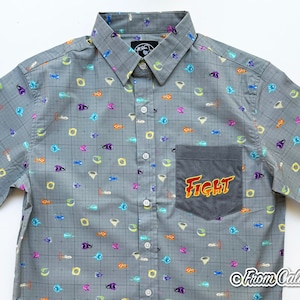 Puede incluir: Camisa gris con botones, con un patr&oacute;n de cuadr&iacute;cula y gr&aacute;ficos de dibujos animados coloridos. La camisa tiene un bolsillo con la palabra "FIGHT" en un dise&ntilde;o de llama. La camisa tiene mangas cortas y un cuello cl&aacute;sico.