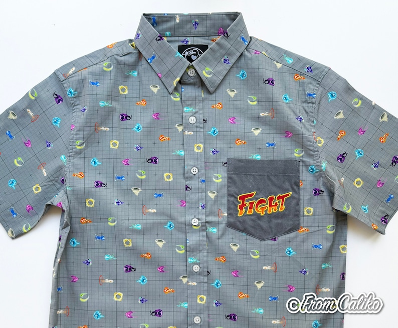 Puede incluir: Camisa gris con botones, con un patr&oacute;n de cuadr&iacute;cula y gr&aacute;ficos de dibujos animados coloridos. La camisa tiene un bolsillo con la palabra "FIGHT" en un dise&ntilde;o de llama. La camisa tiene mangas cortas y un cuello cl&aacute;sico.