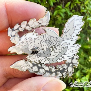 May include: A detailed enamel pin featuring a stylized, white and gray creature with a fierce expression. The pin has a gold outline and is held in a hand. The pin is a collectible item.