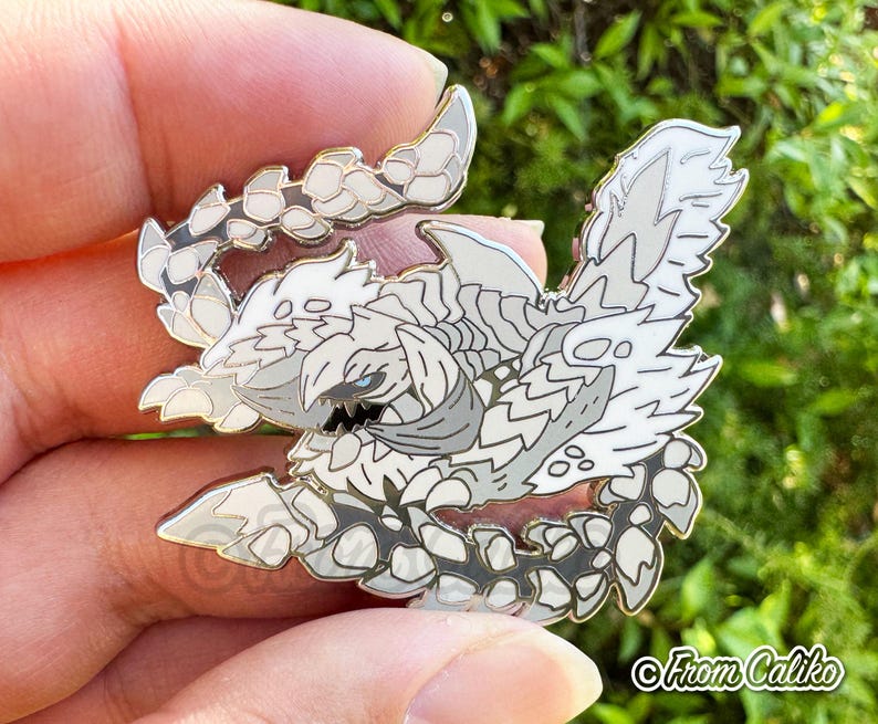 May include: A detailed enamel pin featuring a stylized, white and gray creature with a fierce expression. The pin has a gold outline and is held in a hand. The pin is a collectible item.